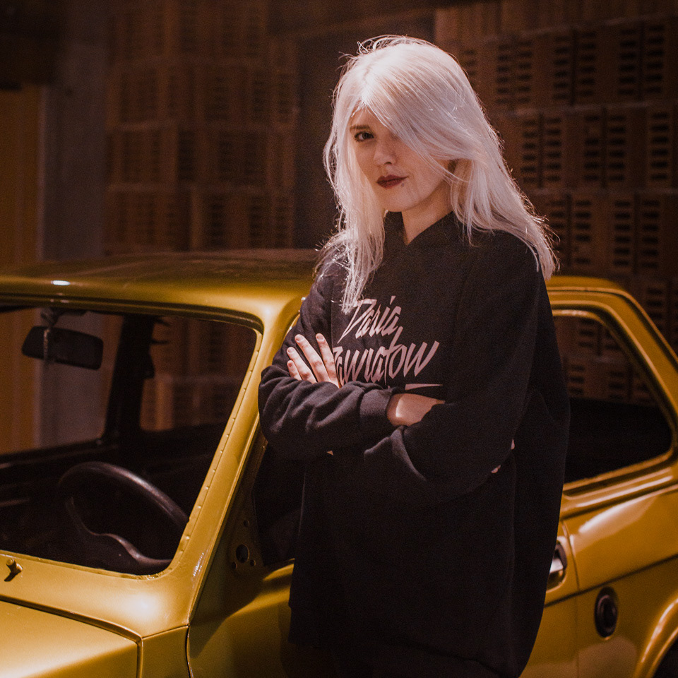 Singer Daria Zawiałow poses next to a shiny, golden Fiat 126p, holding hands (a gesture of friendship/collaboration).