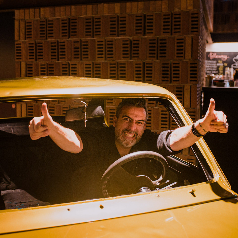 Stand-up comedian Abelard Giza is joyful inside a golden Maluch without a windshield, reaching out to the camera and showing thumbs up.