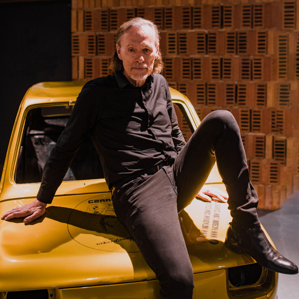 John Porter in a rock pose sits on the hood of the golden car, resting his hands on the bodywork.